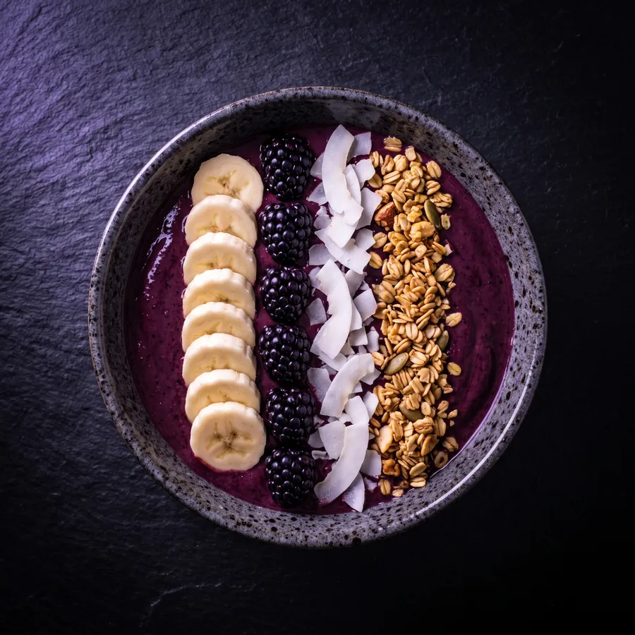 Violette Smoothie-Bowl mit Bananenscheiben, Heidelbeeren und Granola.